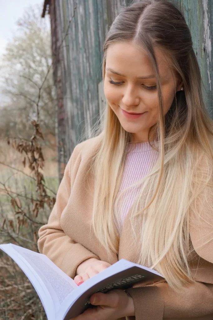 Eine junge Frau lächelt sanft, während sie draußen ein Buch liest.