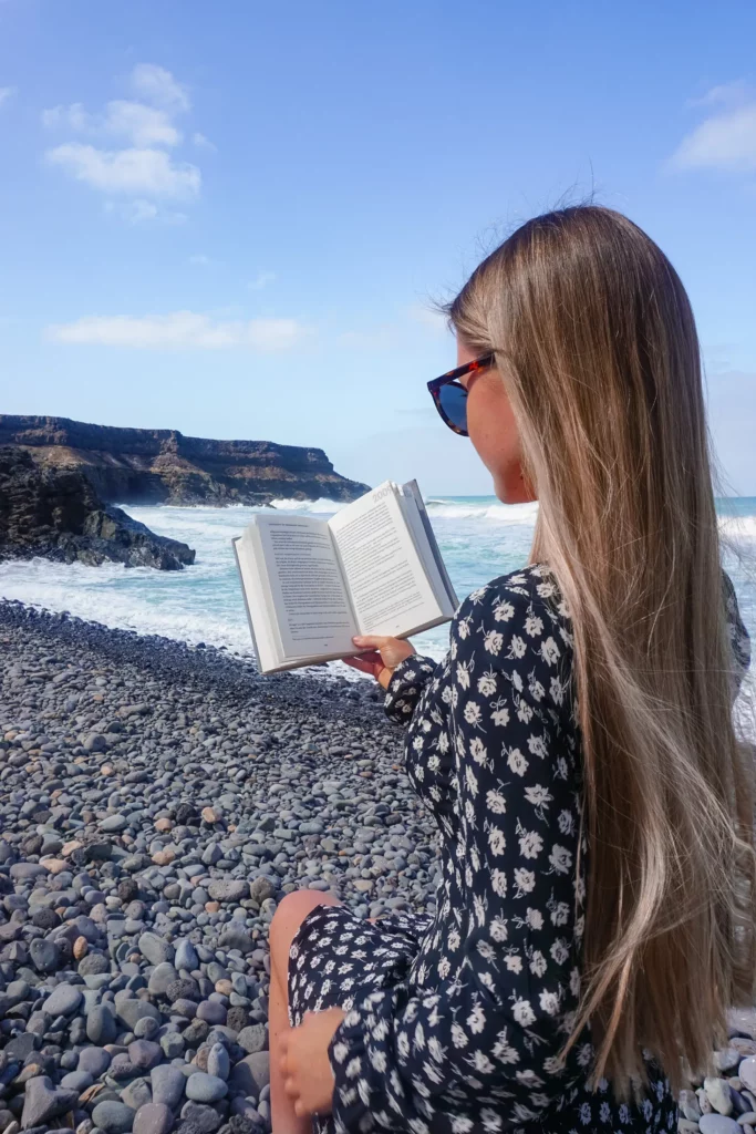 Frau liest ein Buch an einem Kiesstrand, im Hintergrund brechen die Wellen.