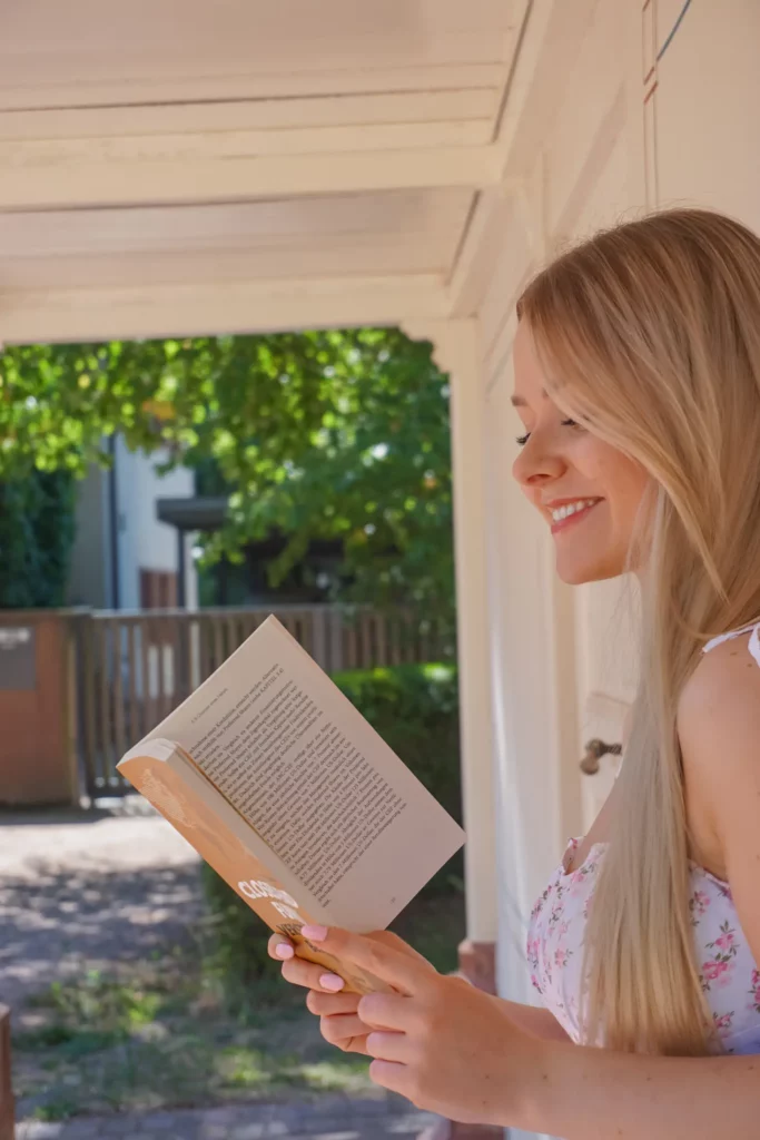 Eine Frau lächelt, während sie draußen unter einer schattigen Veranda ein Buch liest.