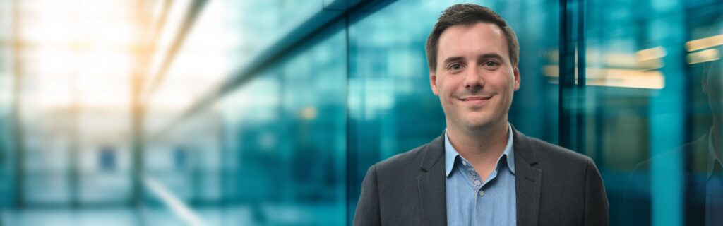 Ein lächelnder Mann im Business-Outfit, Clemens Faustenhammer, steht vor einer gläsernen Gebäudefassade.