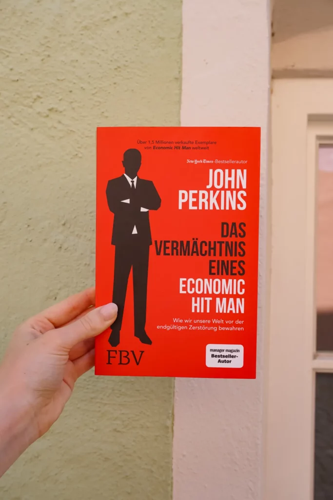 One hand holds a red book entitled "Confessions of an Economic Hitman" by John Perkins in German against a pink wall background.
