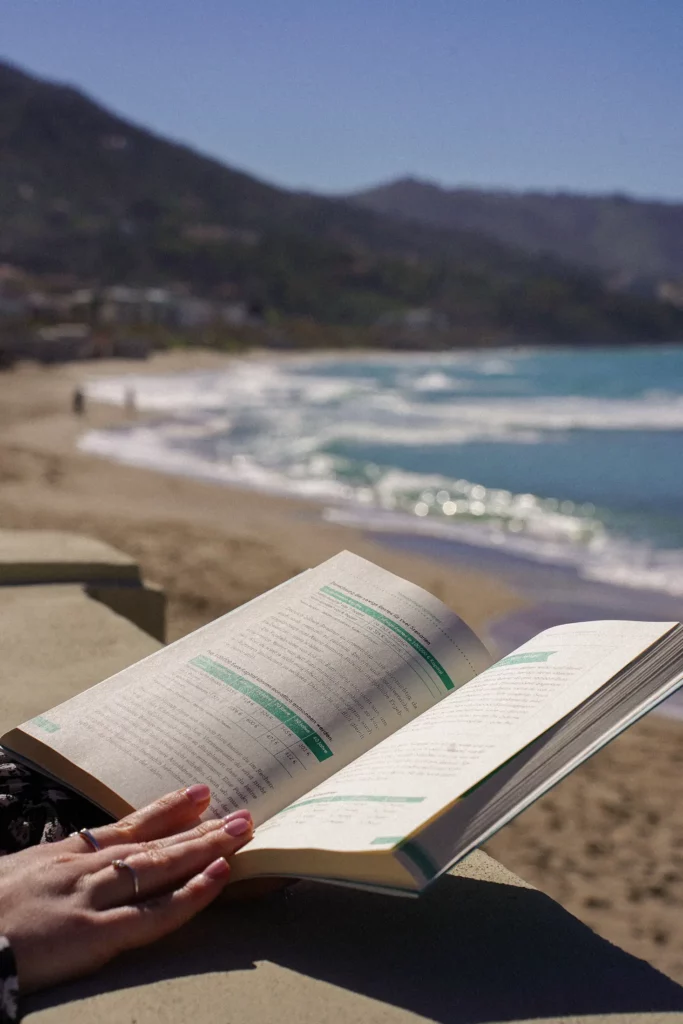 Person liest ein Buch an einem sonnigen Strand mit Wellen im Hintergrund.