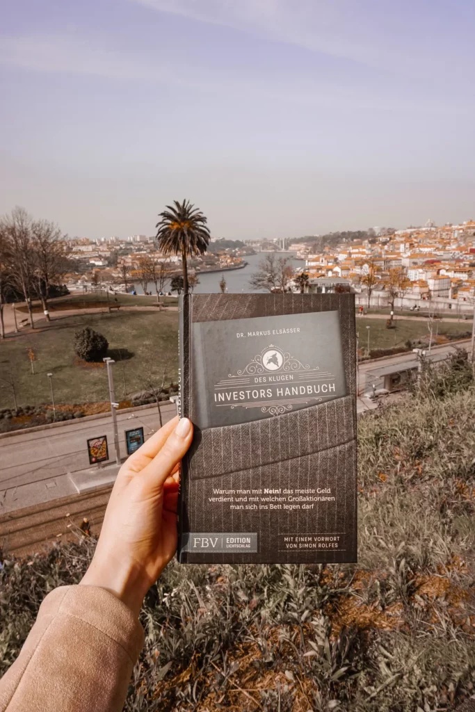 Person hält an einem klaren Tag ein Buch mit dem Titel „Handbuch für Investoren“ mit einer Stadtlandschaft und einem Fluss im Hintergrund.