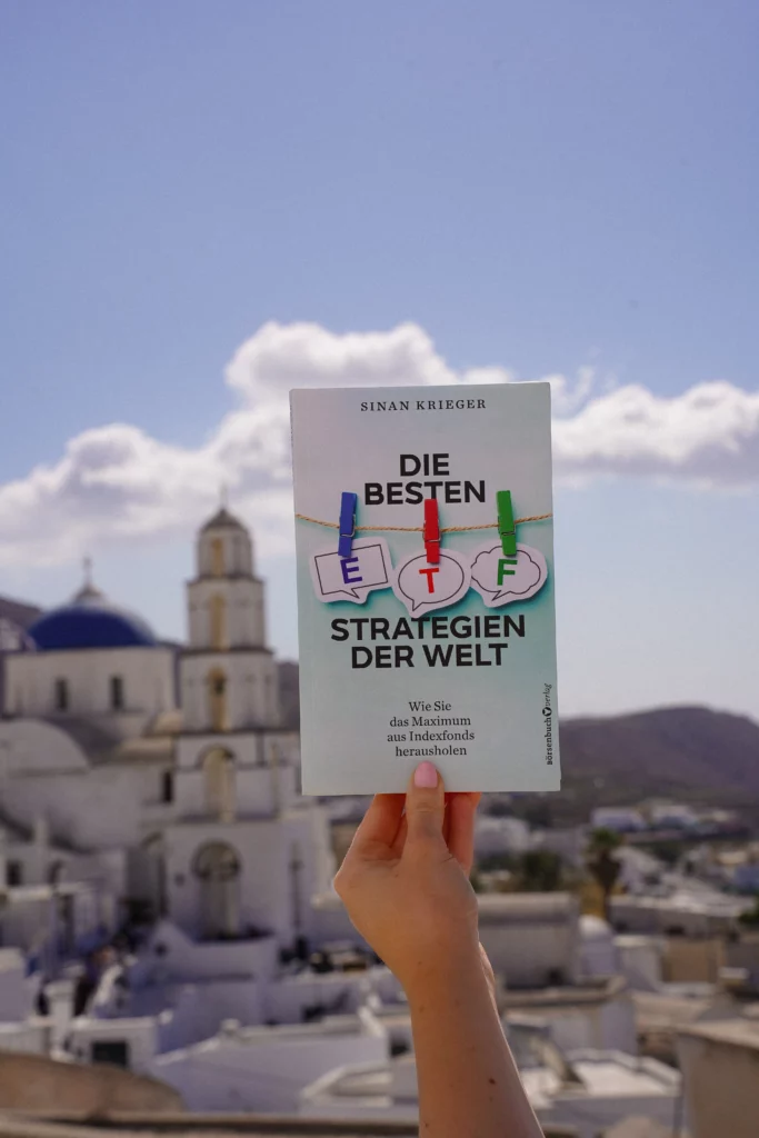 One hand holds a book entitled "The Best ETF Strategies" with a picturesque view of white buildings and blue domes in the background.
