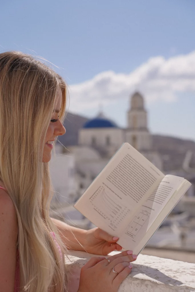 Frau liest ein Buch über ETF-Strategien mit malerischen weißen Gebäuden und einer blauen Kuppel im Hintergrund.