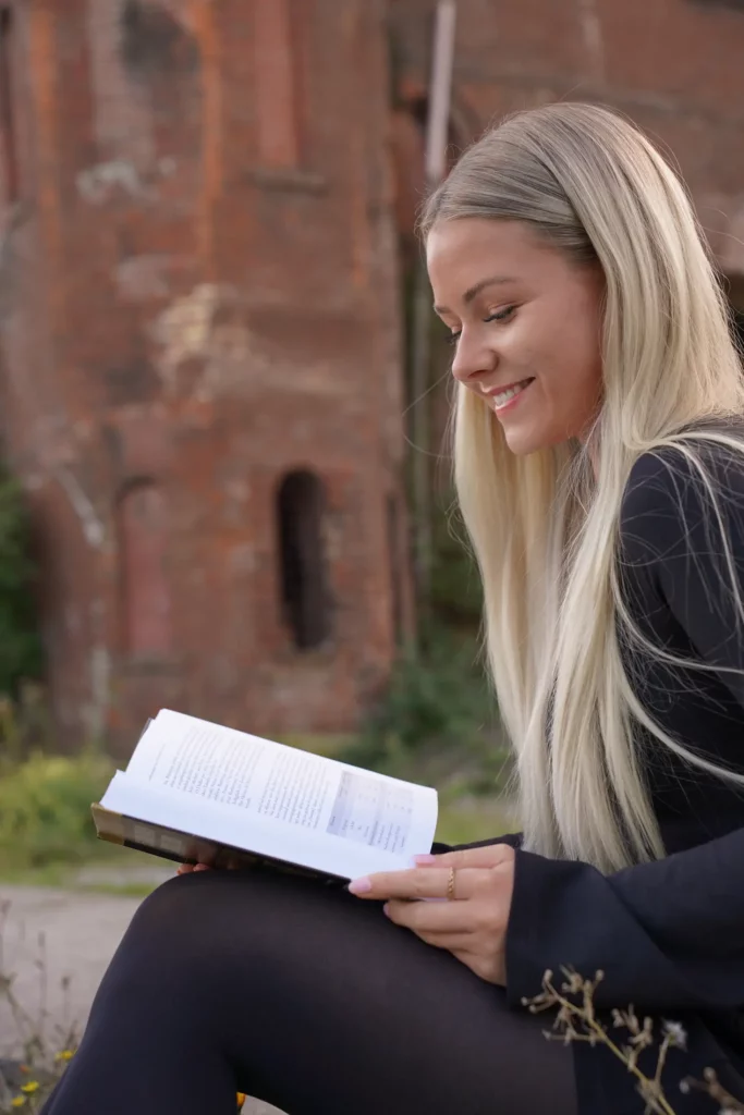 Eine Frau lächelt, während sie draußen ein Buch liest, mit einer Backsteinkonstruktion im Hintergrund.