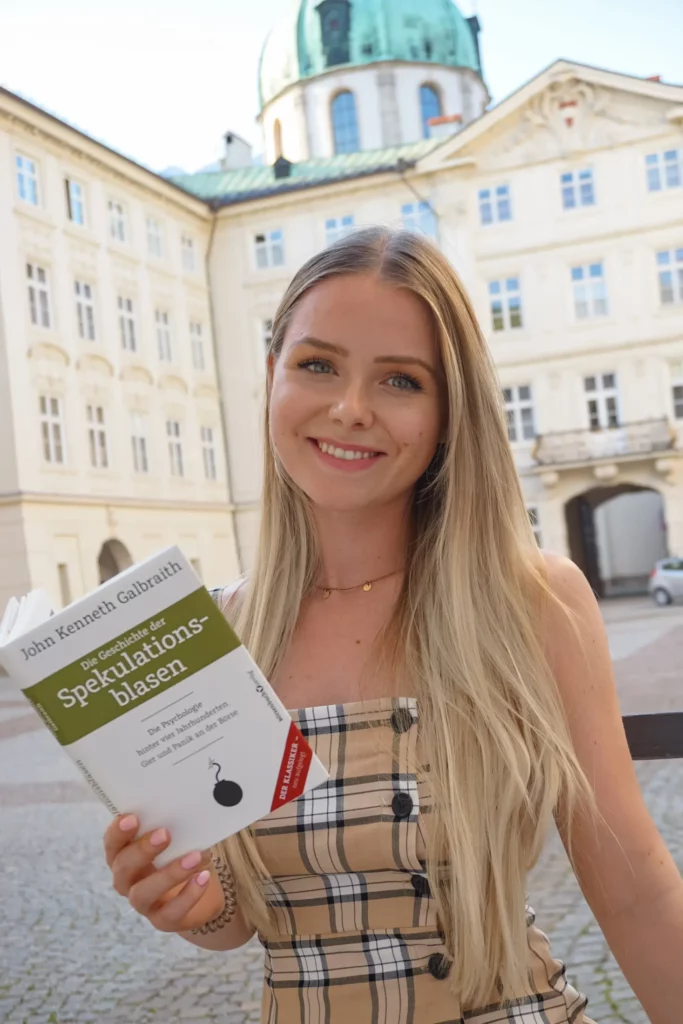 A young woman smiles and holds a book entitled "speculative bubbles" outside near a historic building.