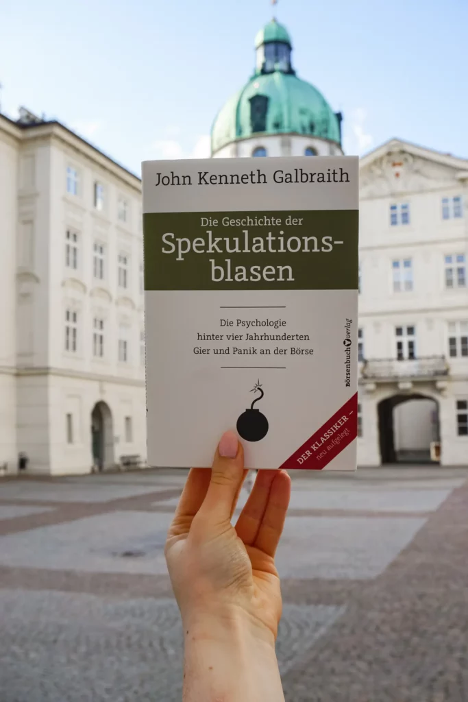 A person holds a copy of John Kenneth Galbraith's book "The History of Speculative Bubbles" in front of a historic building with a green dome.