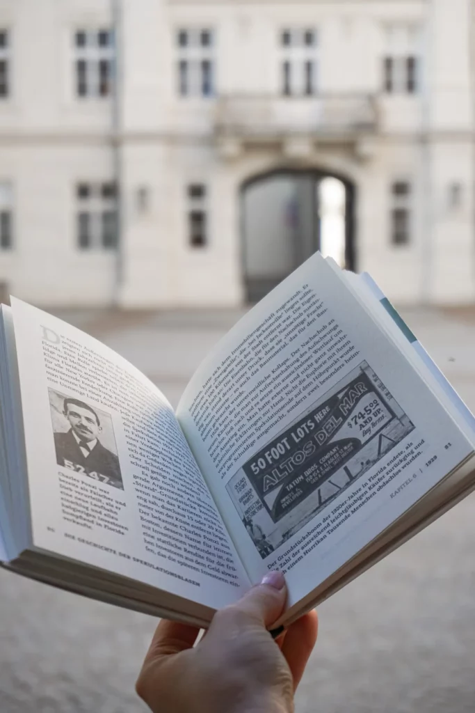 Person holding an open book with text and pictures in front of the blurred background of a building with classical architecture.