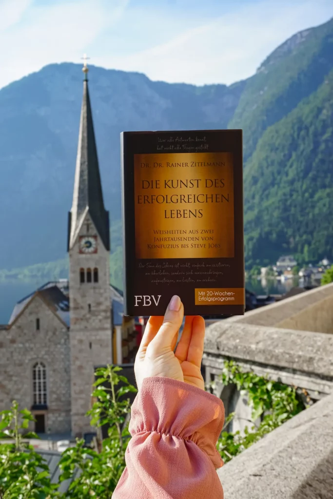 Eine Hand hält ein Buch mit dem Titel „Die Kunst des erfolgreichen Lebens“ hoch, im Hintergrund eine Kirche und Berge.