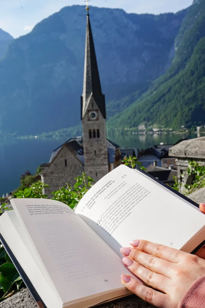 Ein Buch lesen mit Blick auf einen Kirchturm und einen Bergsee im Hintergrund.