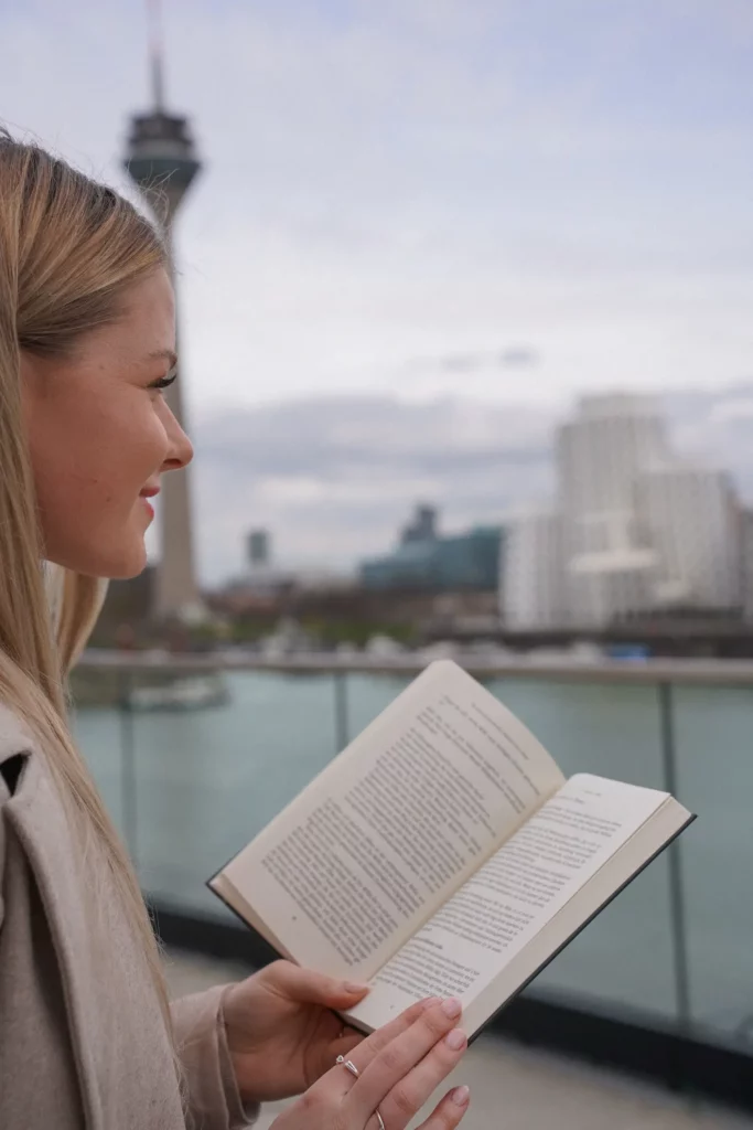 Eine Frau liest ein Buch mit einem städtischen Flussufer und einem Turm im Hintergrund.