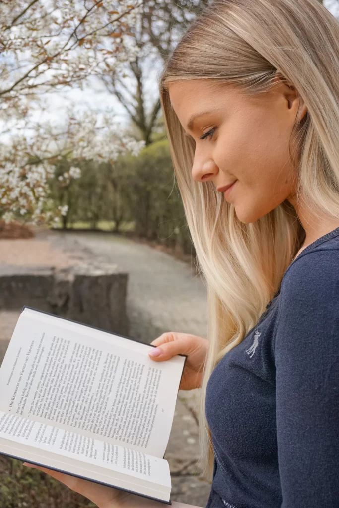 Frau liest draußen ein Buch mit blühenden Bäumen im Hintergrund.
