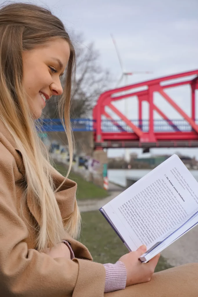Lächelnde Frau beim Lesen eines Buches im Freien mit einer roten Brücke im Hintergrund.