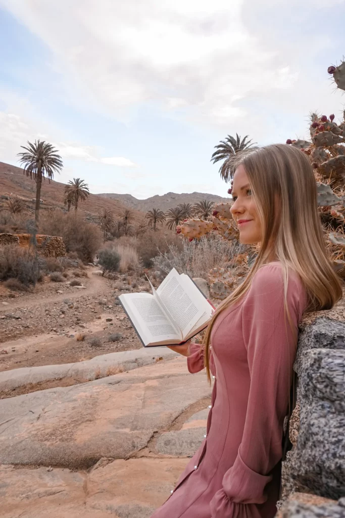 Eine Frau liest ein Buch im Freien in einer wüstenähnlichen Landschaft mit Palmen im Hintergrund.