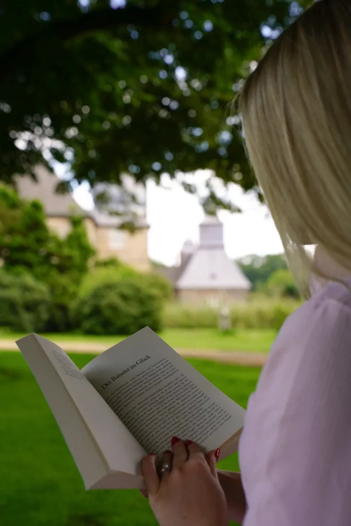 Eine Person liest ein Buch vor einem verschwommenen Hintergrund aus Grünflächen und Gebäuden.
