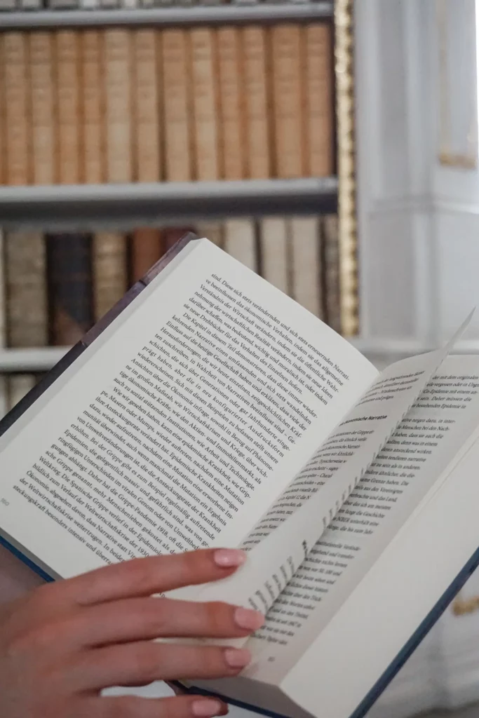 Person hält ein aufgeschlagenes Buch mit flatternden Seiten vor einem Bücherregal.
