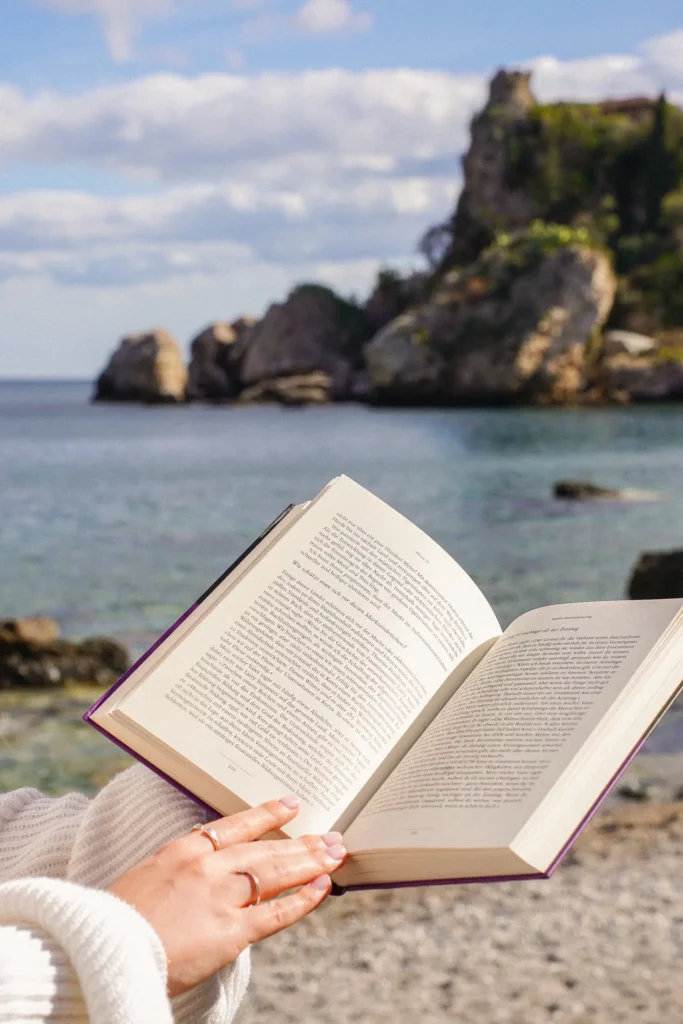 Eine Person liest ein Buch am Meer mit Klippen im Hintergrund.