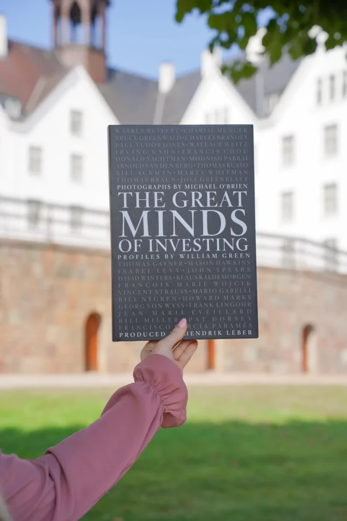 Eine Person hält ein Buch mit dem Titel „The great minds of investing“ vor ein historisches Gebäude und richtet den Buchumschlag an der Fassade des Gebäudes aus.