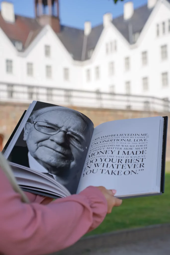 Person hält ein aufgeschlagenes Buch mit einem Schwarz-Weiß-Porträt eines lächelnden Mannes auf der linken Seite und einem sichtbaren Zitat auf der rechten Seite, mit einem verschwommenen Gebäude im Hintergrund.