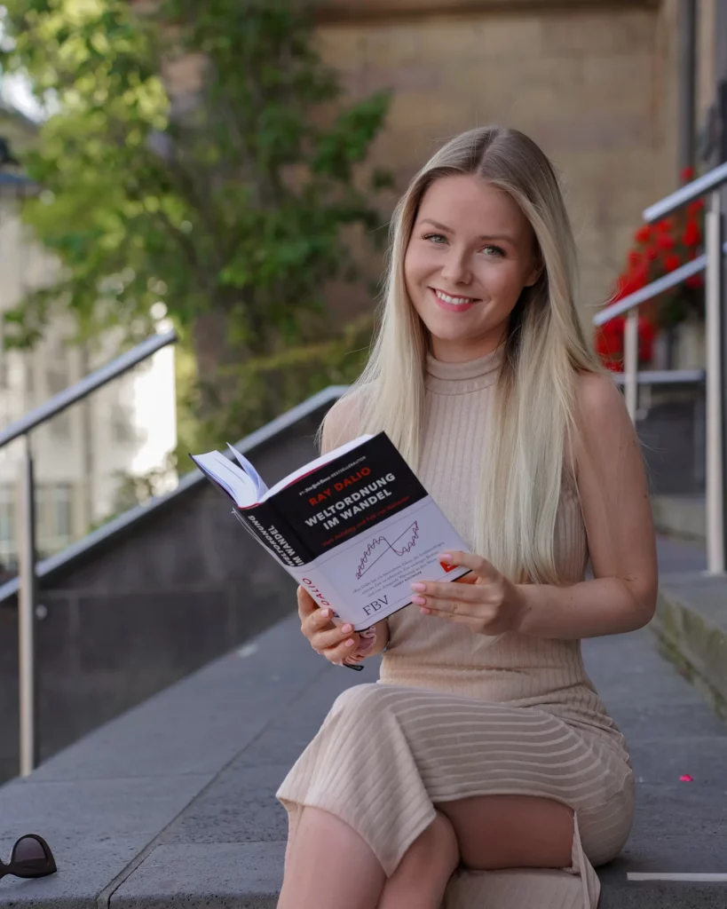 Eine lächelnde Frau sitzt auf einer Treppe und hält ein offenes Buch in der Hand.