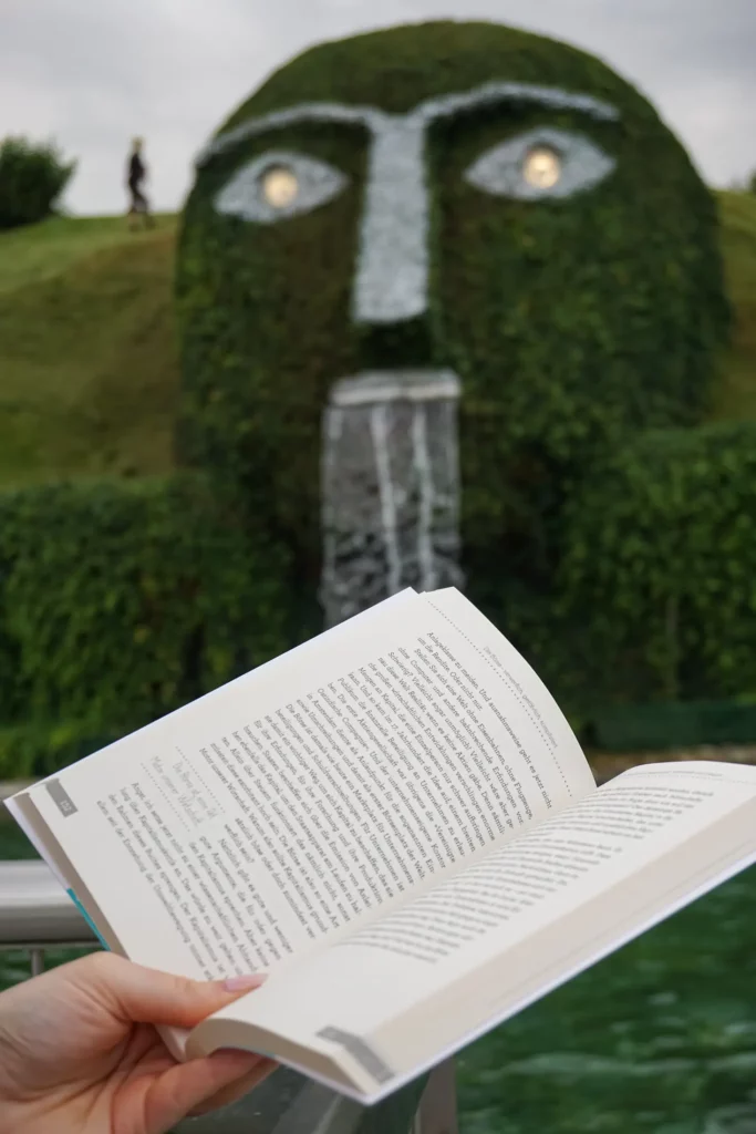 Eine Person hält ein offenes Buch mit einer Brunnenskulptur im Hintergrund, die einem mit Grün bedeckten Gesicht ähnelt.