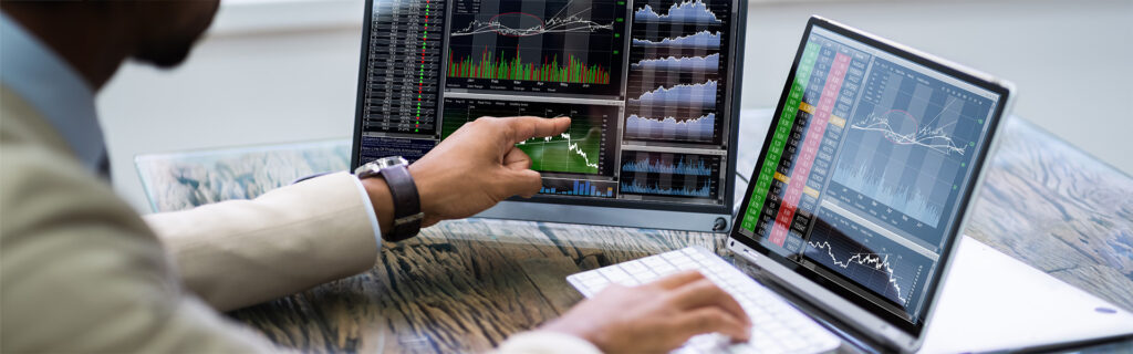 Eine Person in einem Anzug analysiert Finanzcharts und Aktiendaten, einschließlich kurzfristiger Anleihen, die auf zwei Computermonitoren an einem Schreibtisch angezeigt werden.