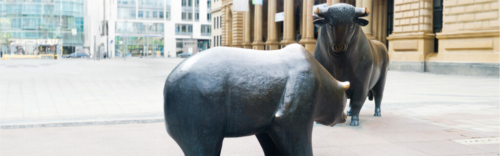 Zwei große Bronzestatuen, bekannt als Bulle und Bär, stehen sich auf einer gepflasterten Fläche gegenüber, die von modernen und historischen Gebäuden umgeben ist.