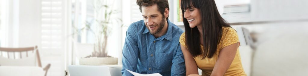 A man and a woman sit together in a house, looking at paperwork and a laptop, looking focused and smiling as they plan to open a securities account.