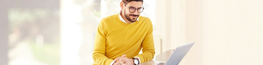 A man with glasses and a yellow sweater is sitting at a desk in a bright room, looking at an open laptop and researching day trading taxes.