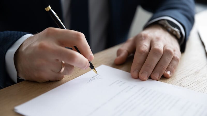 Eine Person im Anzug unterschreibt an einem hölzernen Schreibtisch ein Dokument mit einem Stift und bereitet sich auf "Stiftung gründen" vor.