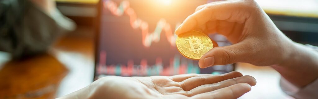 Person, die einer anderen Person einen Bitcoin-Token überreicht, mit einem verschwommenen Börsendiagramm mit ETFs im Hintergrund.