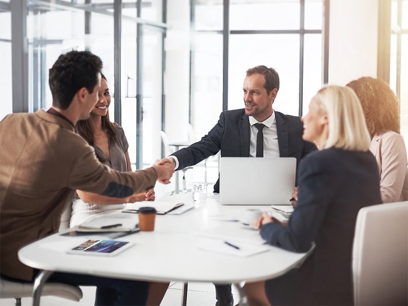 Kollegen in einem modernen Büro sitzen mit Laptops und Dokumenten um einen Tisch und diskutieren über verschiedene Kontoarten. Ein Mann im Anzug schüttelt einem anderen Mann die Hand, wahrscheinlich um einen Bankvertrag abzuschließen.