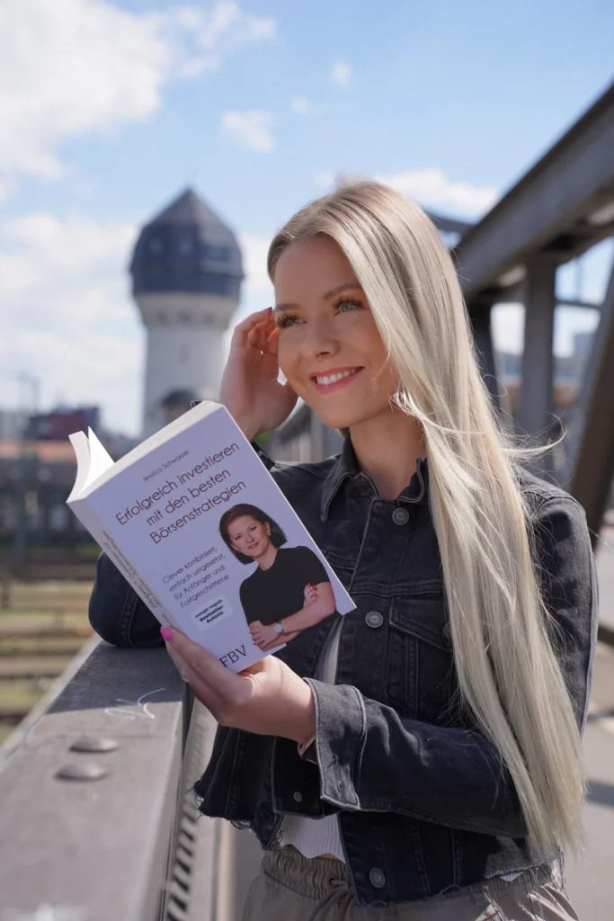 Eine Frau mit langen blonden Haaren und einer Jeansjacke steht im Freien auf einer Brücke und liest ein Buch mit dem Titel „Erfolgreich investieren mit einfachen Börsenstrategien“.