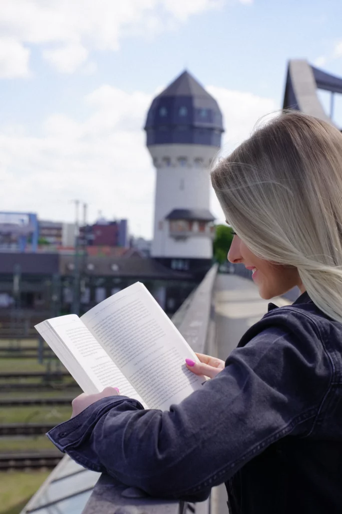Eine Person mit langen Haaren liest draußen auf einer Brücke ein Buch. Im Hintergrund sind ein Turm und Eisenbahnschienen zu sehen.