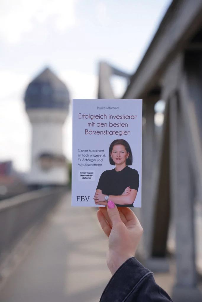 In einer Hand hält man ein Buch mit dem Titel „Erfolgreich Investieren mit den besten Börsenstrategien“ von Jessica Schwarzer. Im Hintergrund ist eine unscharfe Stadtkulisse mit einer Brücke und einem Turm zu sehen.