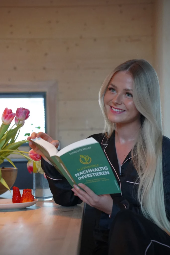 Eine Frau mit blonden Haaren lächelt in die Kamera und hält in einem gemütlichen Holzinterieur ein Buch mit dem Titel „Nachhaltig Investieren“ in der Hand.
