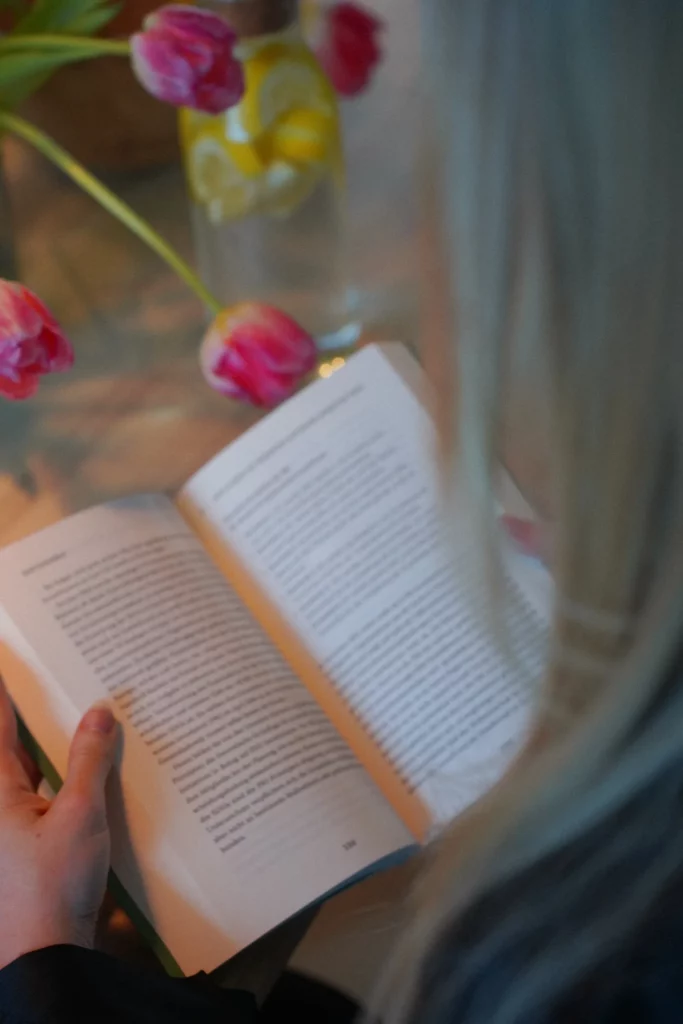 Eine Frau mit blondem Haar liest ein Buch an einem Tisch mit einer Vase mit rosa Tulpen und einem Getränk in weichem Fokus im Hintergrund.