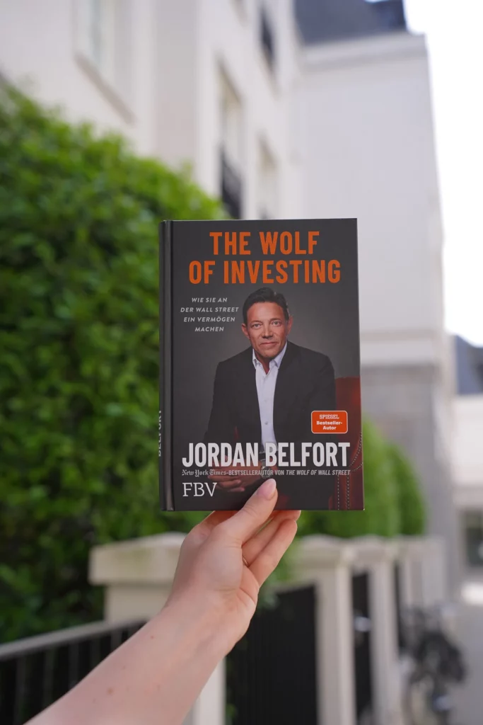Eine Hand hält ein Buch mit dem Titel „The Wolf of Investing“ von Jordan Belfort, abgebildet vor einem unscharfen Gebäudehintergrund.
