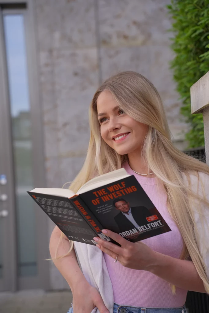 Eine junge Frau lächelt, während sie im Freien das Buch „The Wolf of Wall Street“ liest.