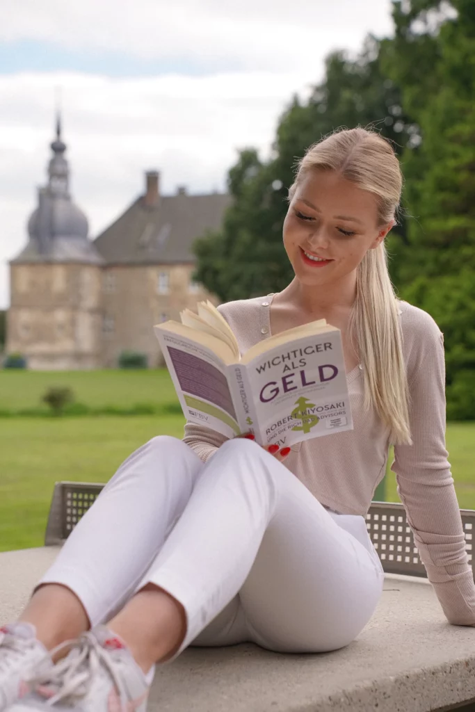 Eine junge Frau sitzt gemütlich im Freien neben einem historischen Gebäude und liest das Buch mit dem Titel „Wichtiger Geld“.