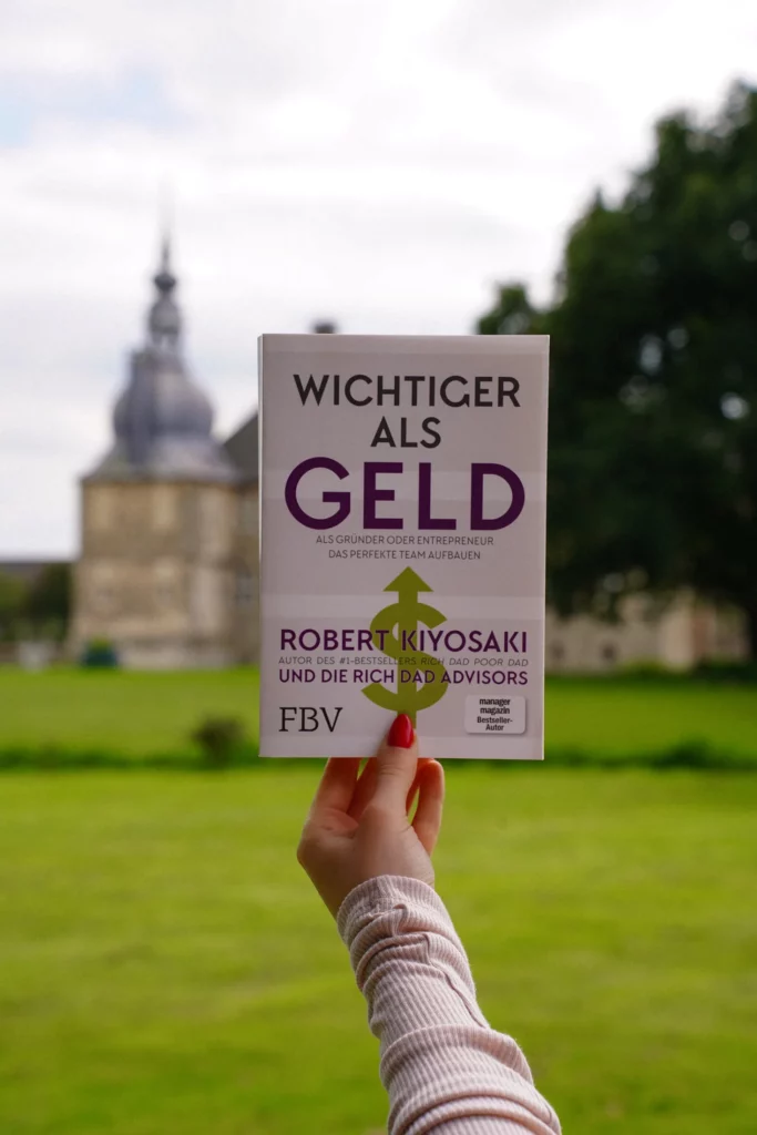 Eine Hand hält ein Buch mit dem Titel „Wichtiger als Geld“ von Robert Kiyosaki vor einem historischen Gebäude mit gepflegtem Rasen hoch.