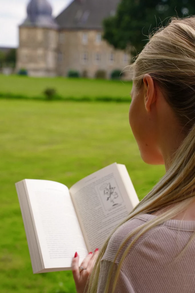 Frau liest im Freien ein Buch mit einem historischen Gebäude im Hintergrund.