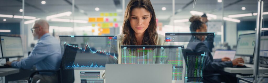 Eine Frau arbeitet an einem Schreibtisch in einem Büro, umgeben von mehreren Monitoren, auf denen Diagramme und Daten angezeigt werden. Im Hintergrund arbeiten weitere Personen an Schreibtischen.