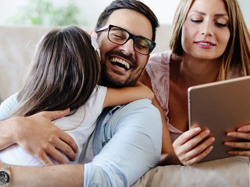 Ein Mann und ein Kind umarmen sich freudig auf einem Sofa, während eine Frau neben ihnen sitzt und sich mit ihrem Tablet beschäftigt und verschiedene Kontotypen recherchiert.