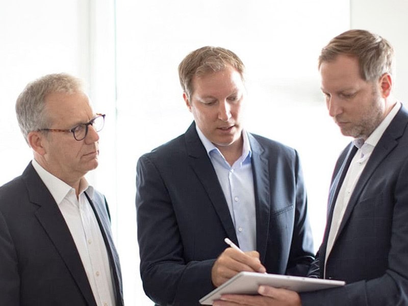 Die Geschäftsführer Michael Heyder, Christian weiß und Andreas Weiß betrachten ein Dokument. Christian Weiß hält einen Stift in der Hand und scheint den anderen etwas über verschiedene Kontotypen zu erklären.