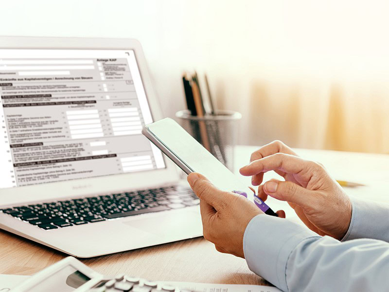 A person is sitting at a desk with a smartphone on which a tax form is displayed. A calculator and some pens can be seen nearby.