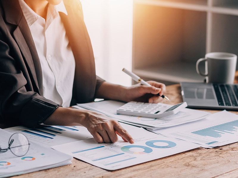 A person in a suit is working at a desk with papers, charts, a calculator and a coffee cup. She points to a circular chart on one of the documents, probably analyzing tax-related data for accurate reporting.