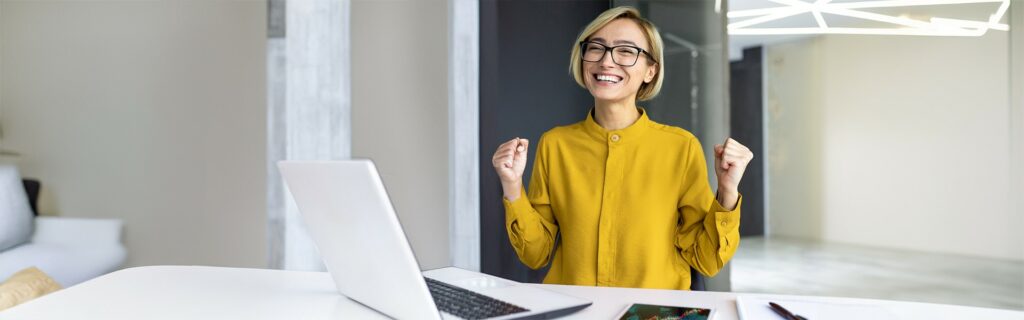 A person with glasses and a yellow shirt is sitting at a desk with a laptop, smiling and raising his fists in excitement.
