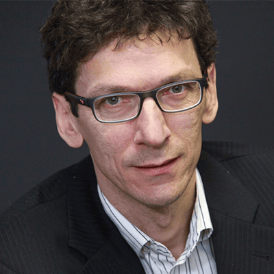 Referent Urban Jaekle mit kurzen dunklen Haaren, Brille und gestreiftem Hemd unter einem dunklen Blazer, der vor einem dunklen Hintergrund in die Kamera blickt und das Selbstvertrauen eines erfahrenen Referenten ausstrahlt.